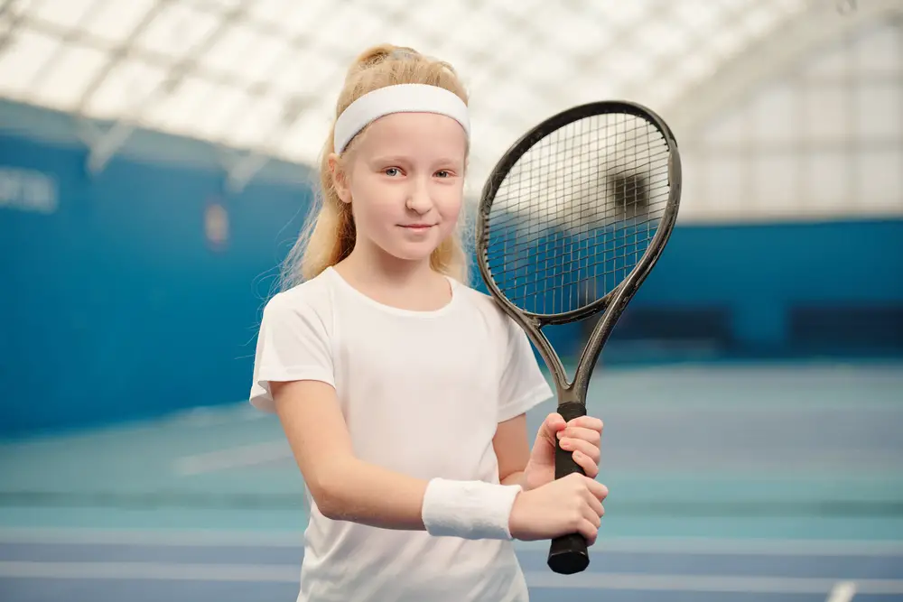 Blond little girl in white activewear holding tennis racket.