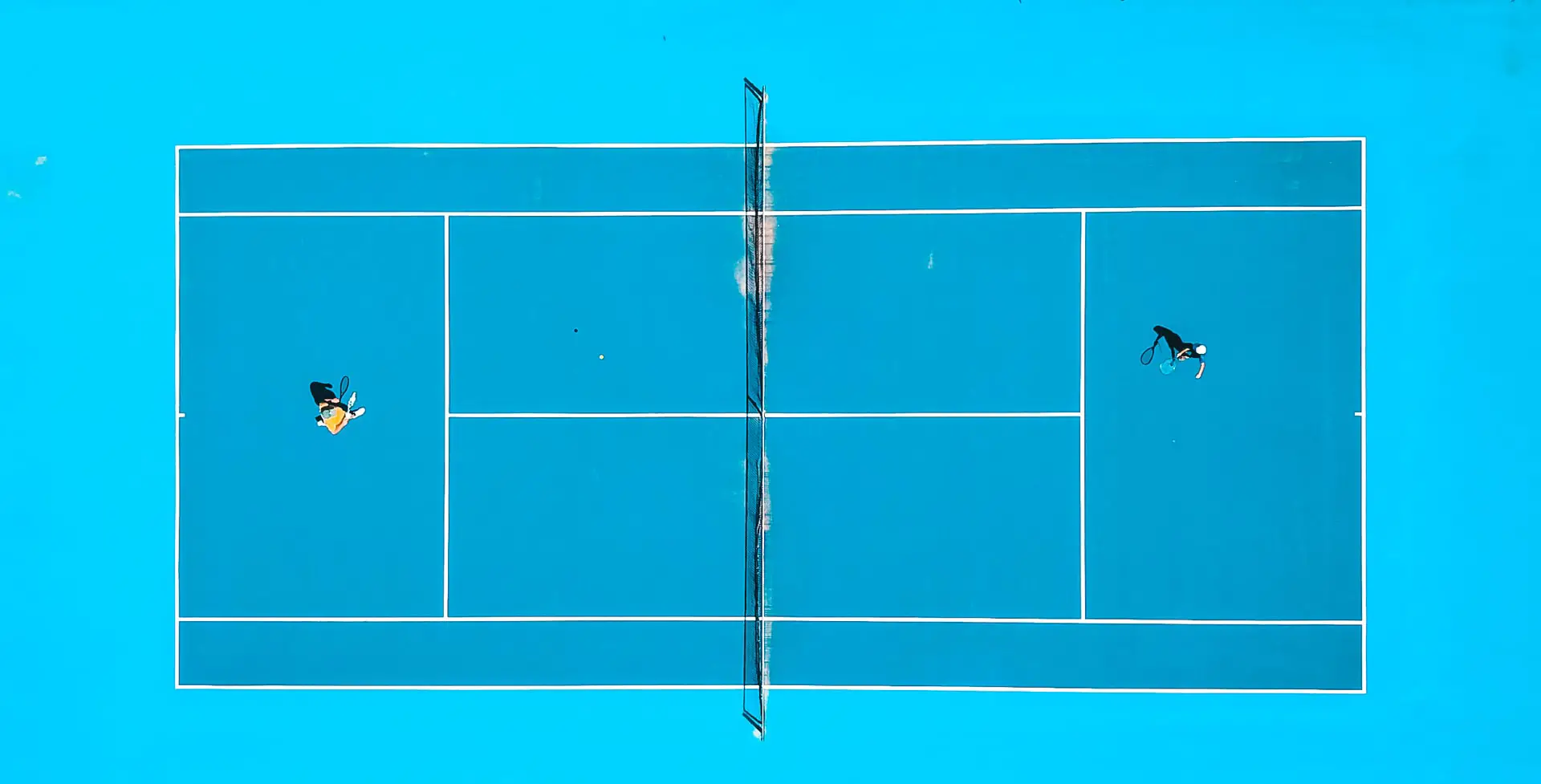 A blue tennis court from bird perspective with two players playing down the line
