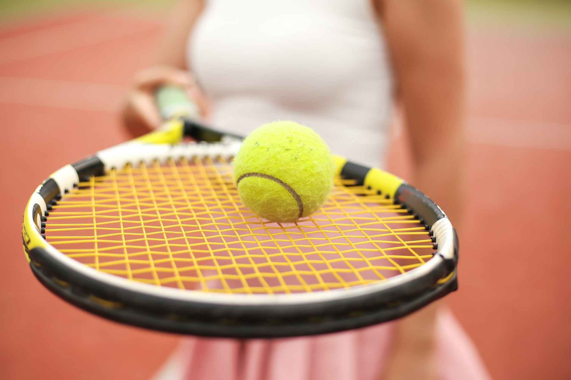 Zoom in of tennis racket with a tennis ball held by a female tennis player in the blurry background